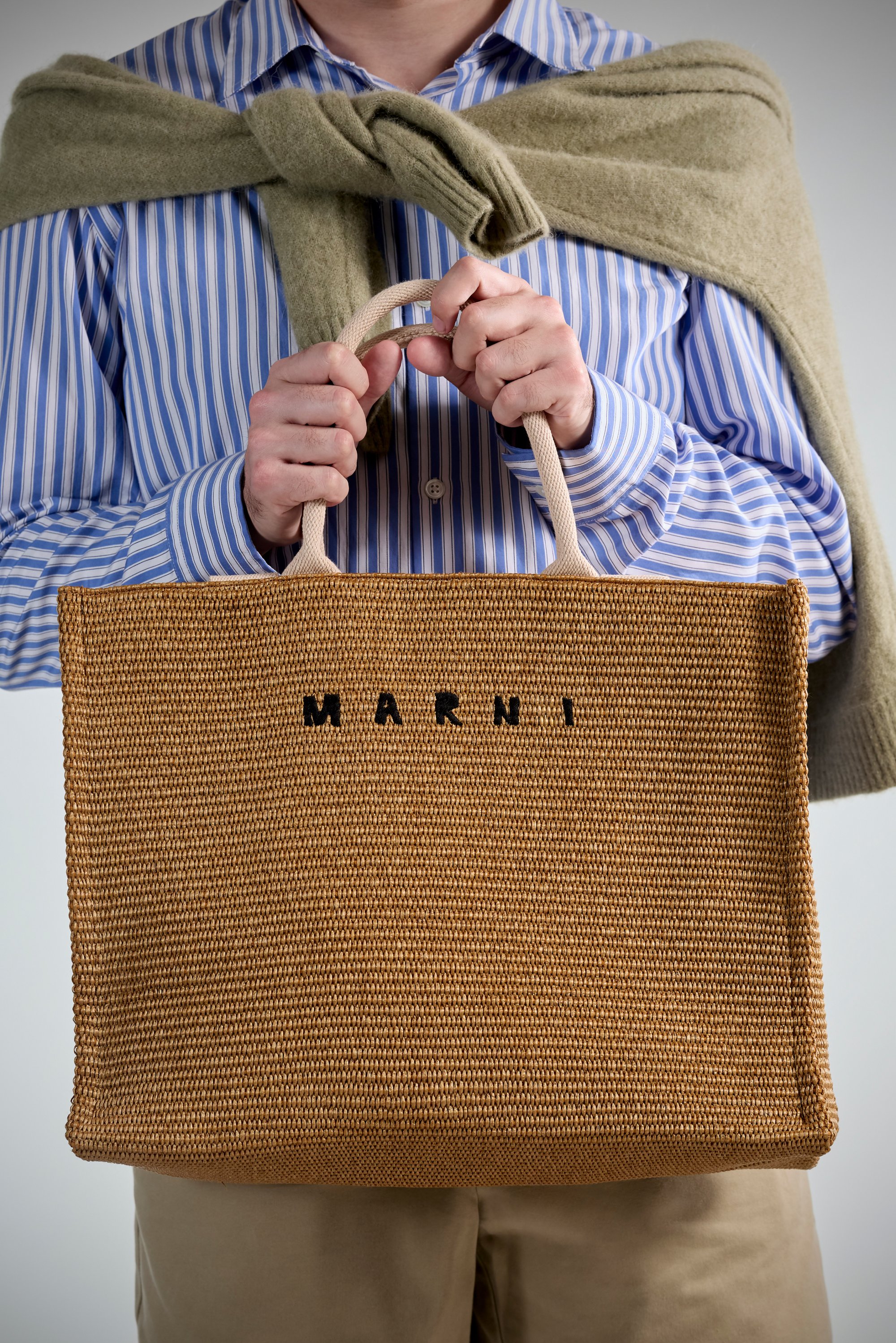 A person holds a woven tan tote bag with "MARNI" printed in black, wearing a blue-striped shirt and olive-green sweater draped over shoulders.