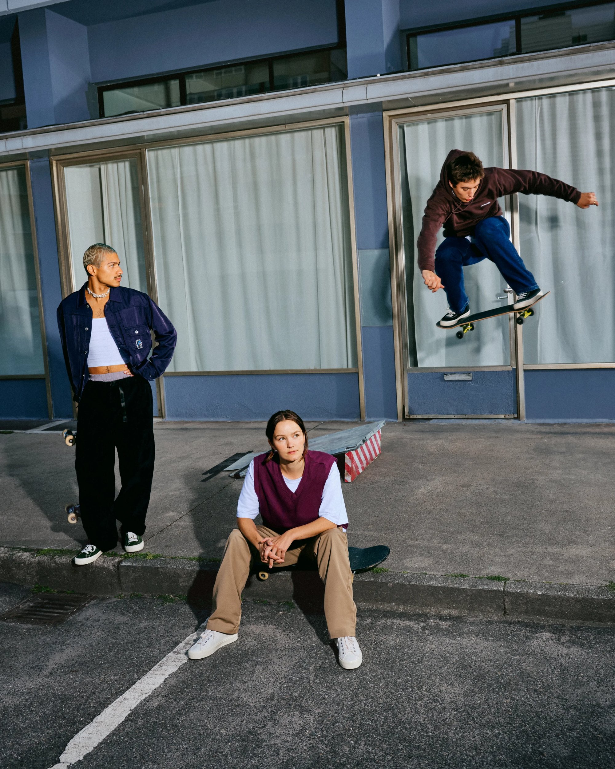 Tre skater in un ambiente urbano: uno esegue un trucco, mentre gli altri due si rilassano. Sono visibili skateboard e rampe, con i riflessi degli edifici sullo sfondo.