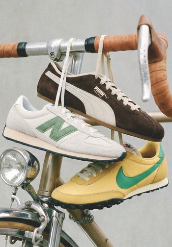 Drie hangende sneakers: een bruine Puma, een witte New Balance met groene "N" en een gele Nike met een groene swoosh, aan een vintage fietsstuur.