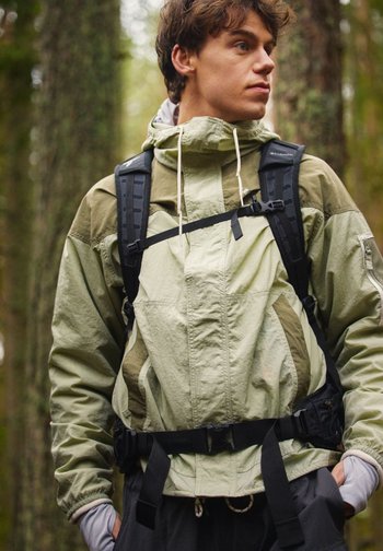 Ung mann med grønn turjakke og svart ryggsekk ser til siden i en skog med høye trær.