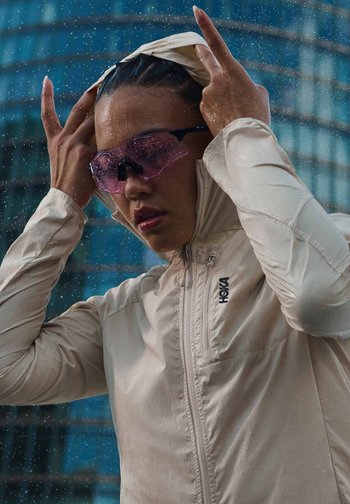 Person wearing light-colored rain jacket and pink tinted sports sunglasses adjusts hood in rainy urban setting.