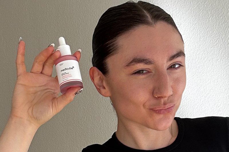Personne aux cheveux plaqués en arrière tient une bouteille rose de sérum pour la peau Medicube près de son visage, portant une chemise noire.