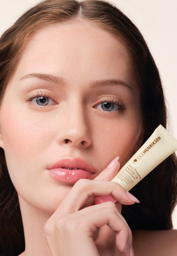 Cream tube with a yellow body and gold text labeled "OLEHENRIKSEN," held near a face with smooth skin and glossy pink lips.