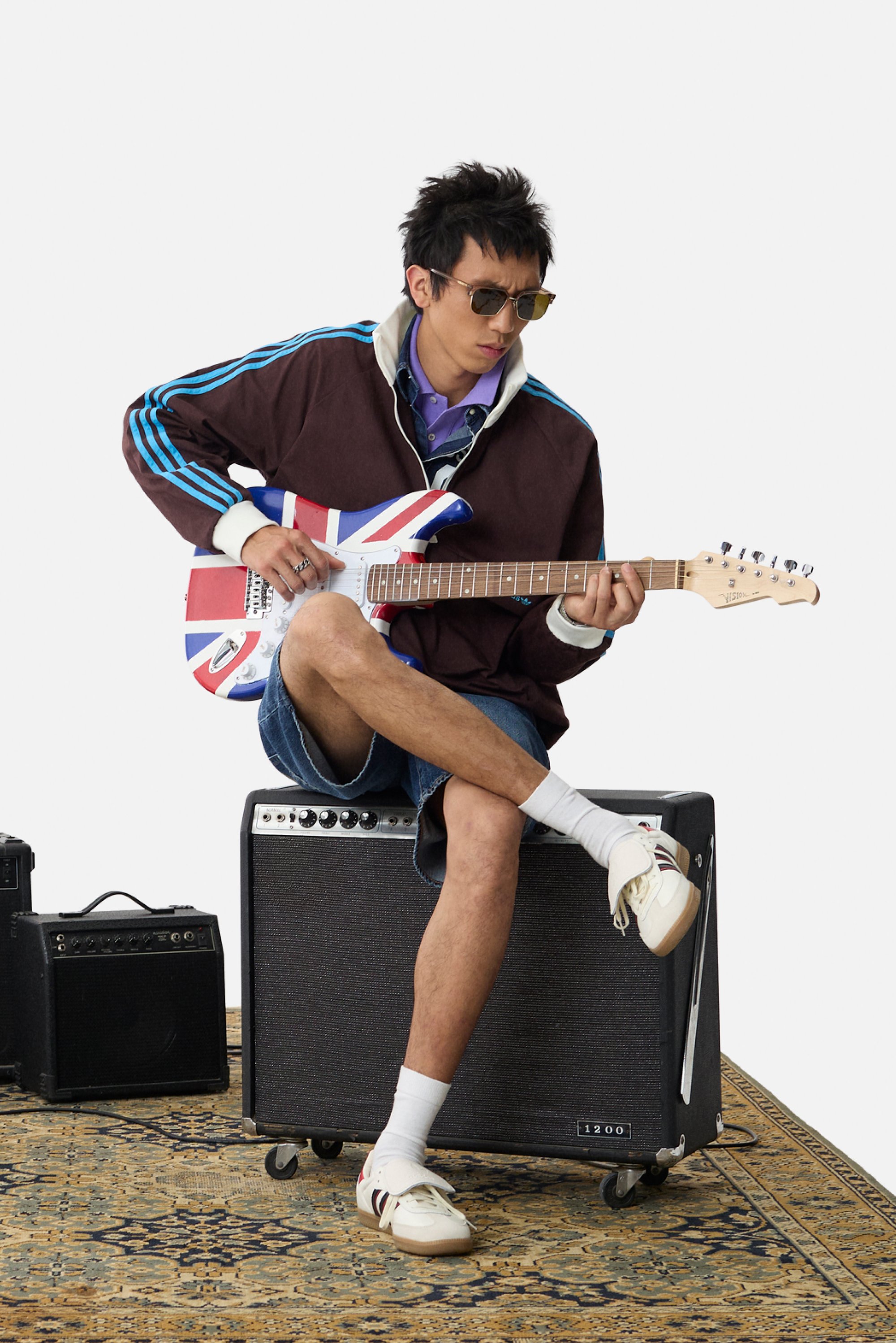 Guitarra eléctrica con diseño de la Union Jack, chaqueta marrón con rayas azules, pantalones cortos de mezclilla, zapatos crema y amplificadores sobre una alfombra con patrones.