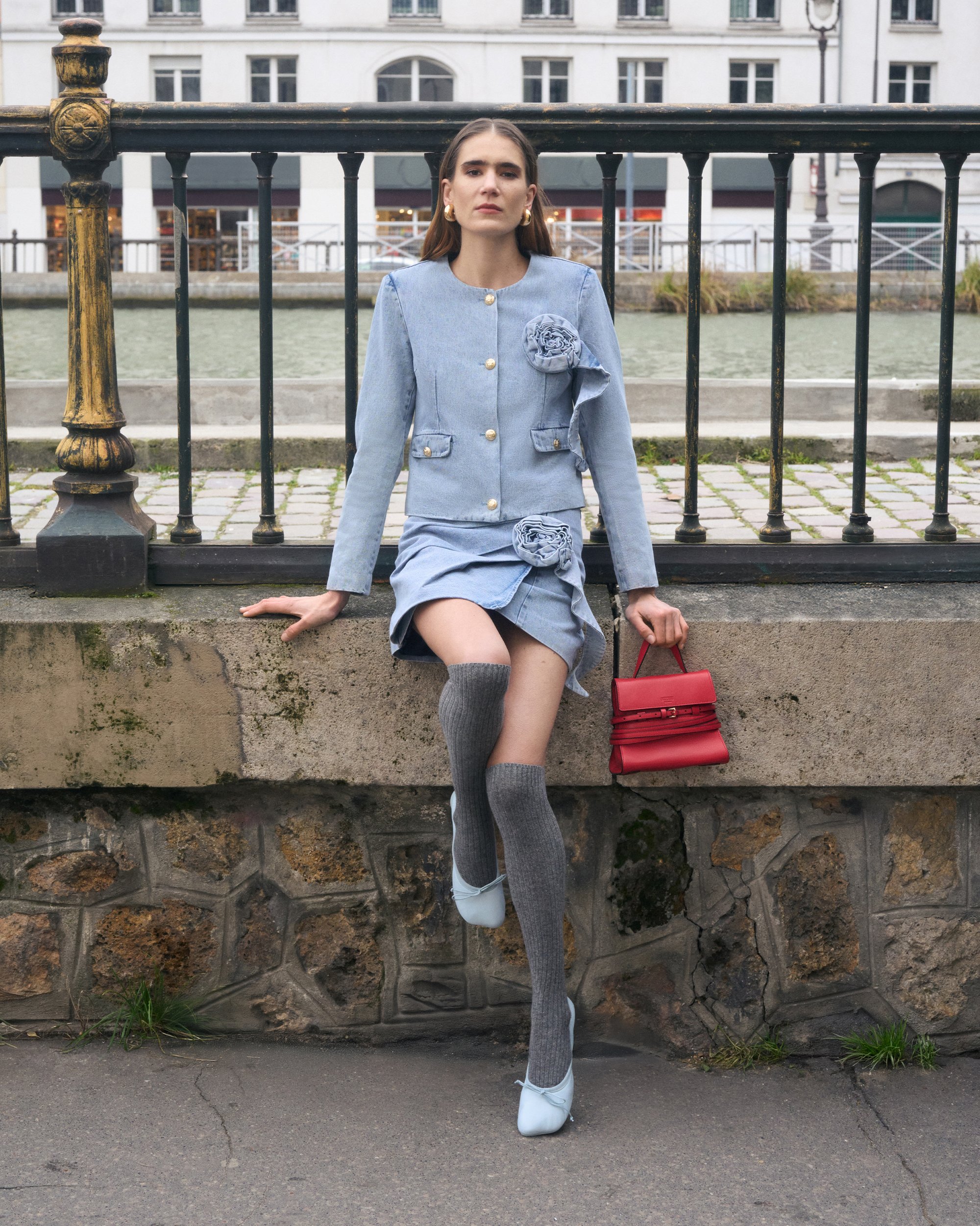 Una modelo se sienta en un muro de piedra, vestida con una chaqueta y una falda azul claro con detalles de flores, combinadas con calcetas grises hasta la rodilla y zapatos azules, sosteniendo un bolso rojo.