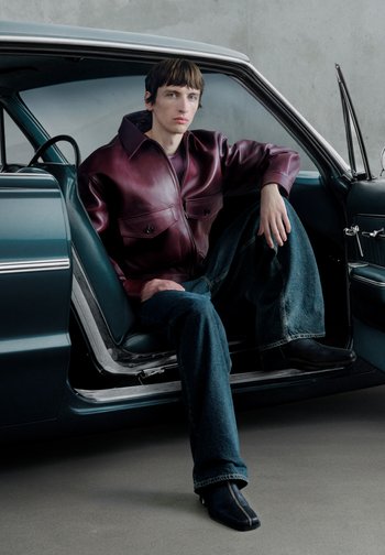 Veste en cuir bordeaux à coupe décontractée, avec deux poches avant ; associée à un jean en denim foncé et des bottines noires à fermeture éclair, assis dans une voiture.