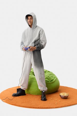 Felpa grigia con cappuccio abbinata a pantaloni, tessuto testurizzato e lacci. Stivali neri, sacco a pelo verde e tappeto rotondo arancione. Controller di gioco in mano.