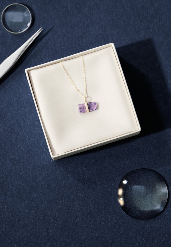 Gold necklace with a chunky purple stone pendant, displayed in a cream box on a dark textured surface; a magnifying glass is placed beside it.