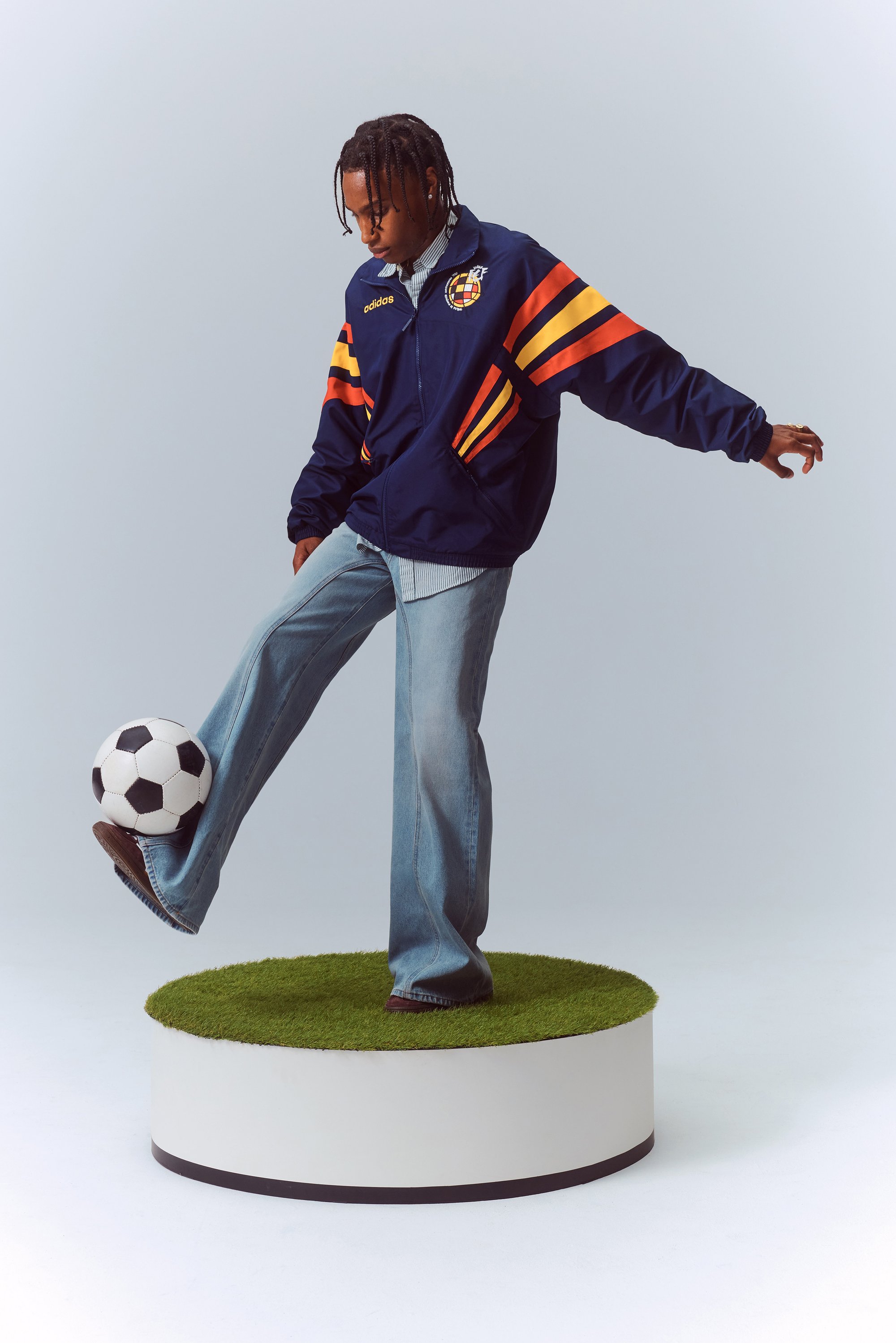 Persona con una giacca blu adidas e pantaloni in denim che bilancia un pallone da calcio sul suo piede, in piedi su un pedestallo circolare coperto di erba.