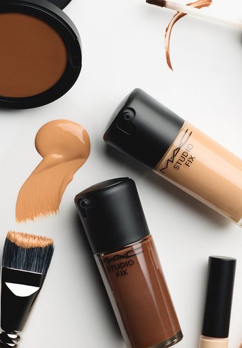 Twee flesjes MAC Studio Fix foundation, een compact met bruine poeder, een borstel met beige foundation en foundation-stalen op een witte ondergrond.