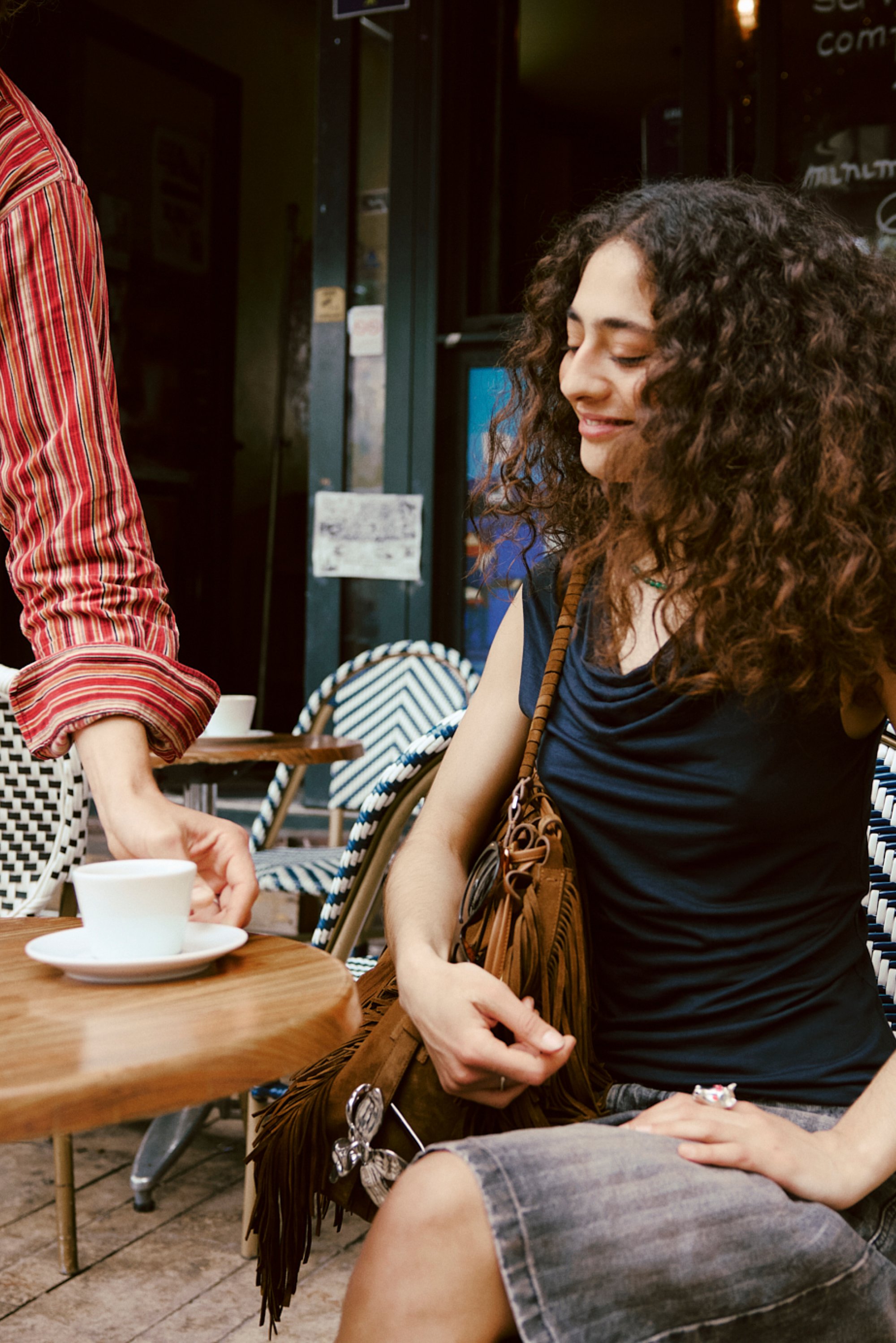 A waiter serves a white coffee cup to a smiling woman with curly hair, seated at an outdoor café table surrounded by patterned chairs.