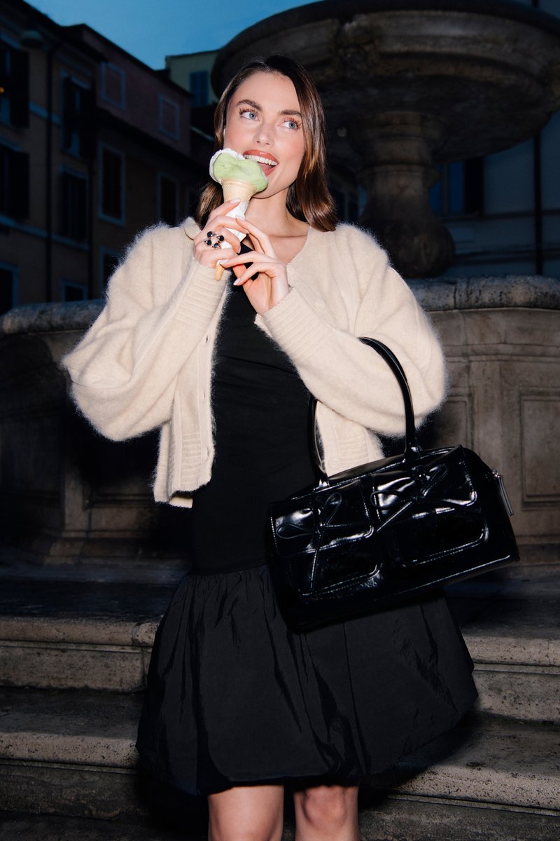 Junge Frau trägt cremefarbene Strickjacke und schwarzes Kleid, hält schwarze Handtasche und isst nachts grünes Eis in der Waffel an einem steinernen Brunnen.