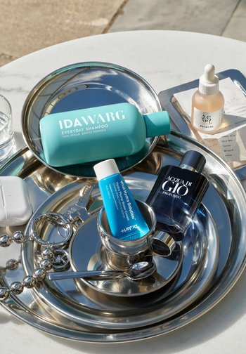 Zilveren schalen bevatten een turquoise shampoo fles, een blauwe crèmetube, een zwarte parfumfles, een transparante druppelfles, een zilveren beker, een lepel, sleutels en een witte doos.