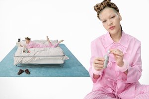 A person in pink striped pajamas applies skincare in a cozy bedroom setting. They're lounging on a bed with beauty products nearby.