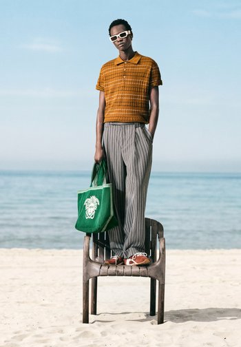 Persoon met witte zonnebril, oranje shirt met korte mouwen, gestreepte broek, bruine schoenen, een groene draagtas vasthoudend, staand op een plastic stoel op een zandstrand.