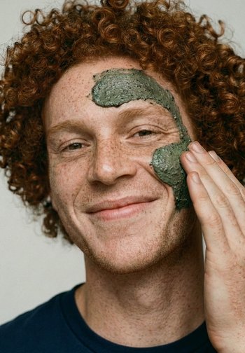 Rødhåret mand med fregner påfører grøn ansigtsmaske på kinden, smilende blidt iført en mørkeblå skjorte mod en ensfarvet baggrund.