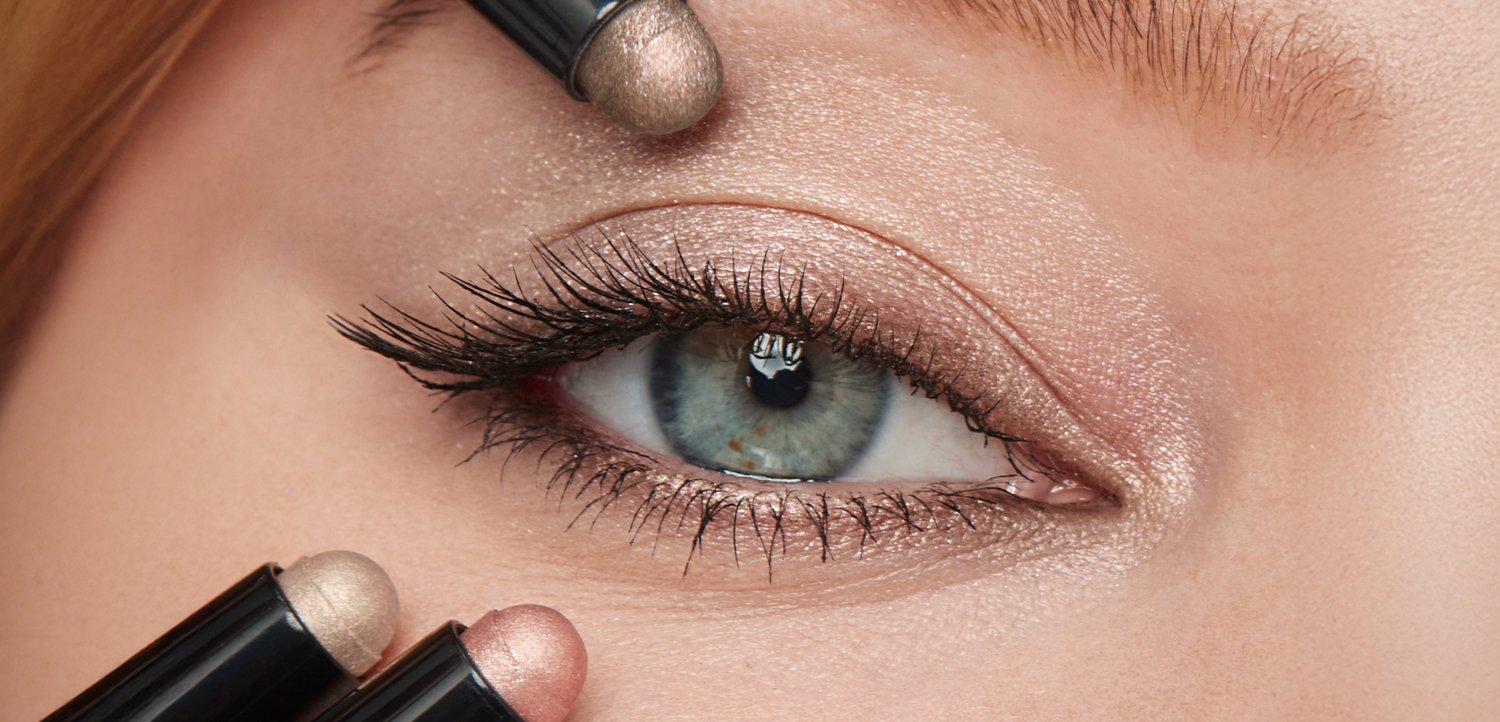Primo piano di un occhio con iris blu-verde, ciglia lunghe e ombretto neutro scintillante applicato con bastoncini per il trucco.
