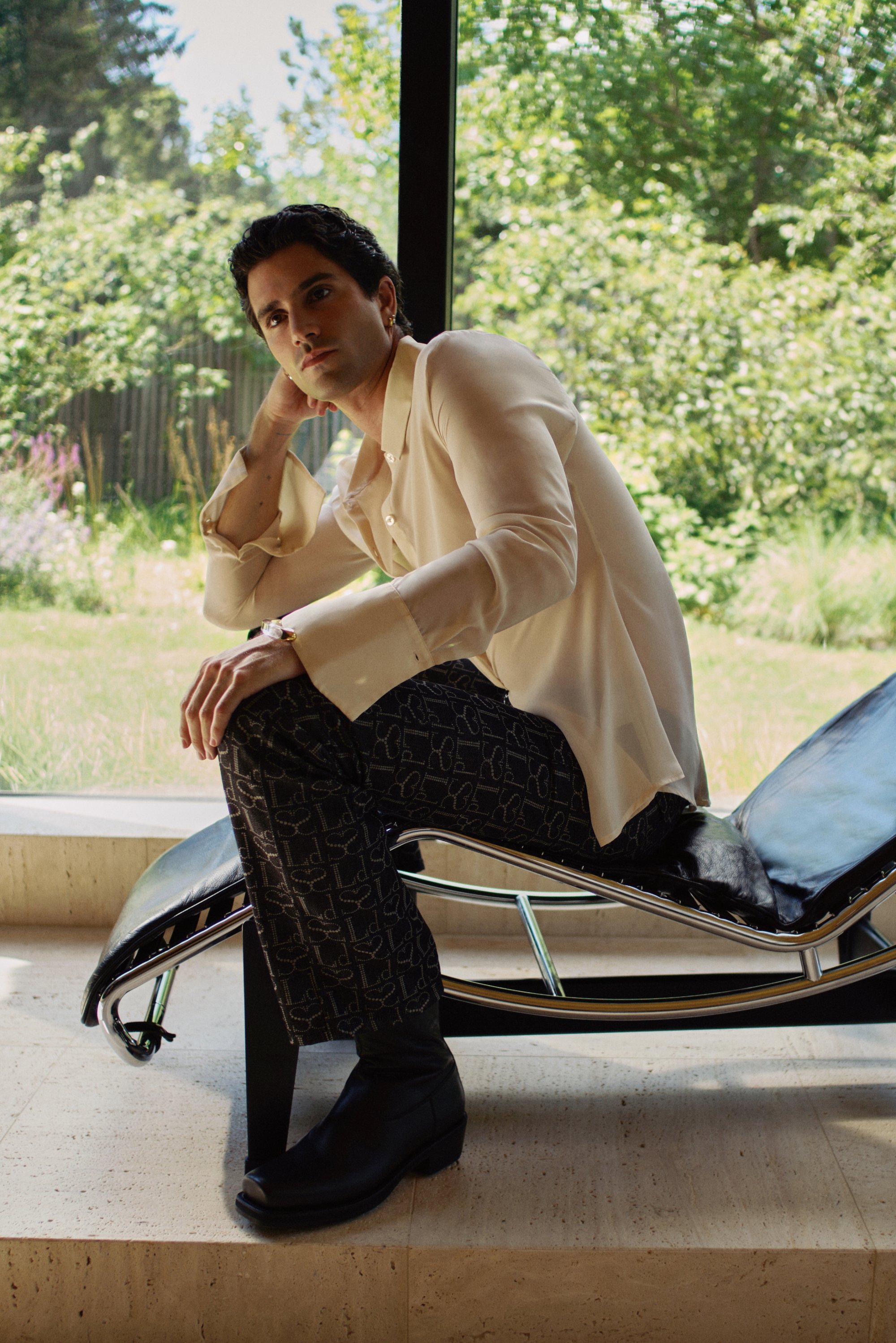 A person in a light shirt and patterned trousers sits on a modern chair, resting their chin on one hand, with lush greenery visible outside.