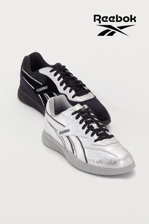 Sort sneakers med fløjl og skinnende accenter sammen med en sølv sneaker med en krøllet tekstur, begge med sorte snørebånd og branding.