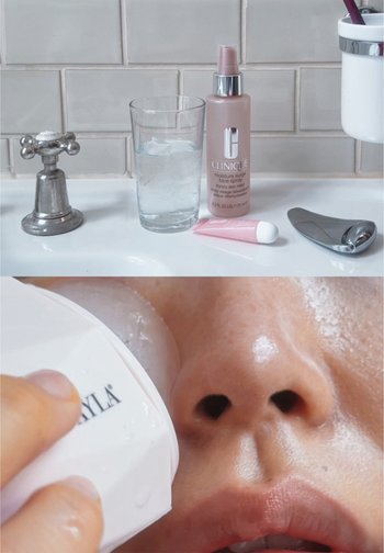 Vaso con agua, botella de spray rosa de Clinique, tubo de crema rosa y una herramienta de masaje plateada sobre una superficie blanca. Primer plano de la piel con el dispositivo.