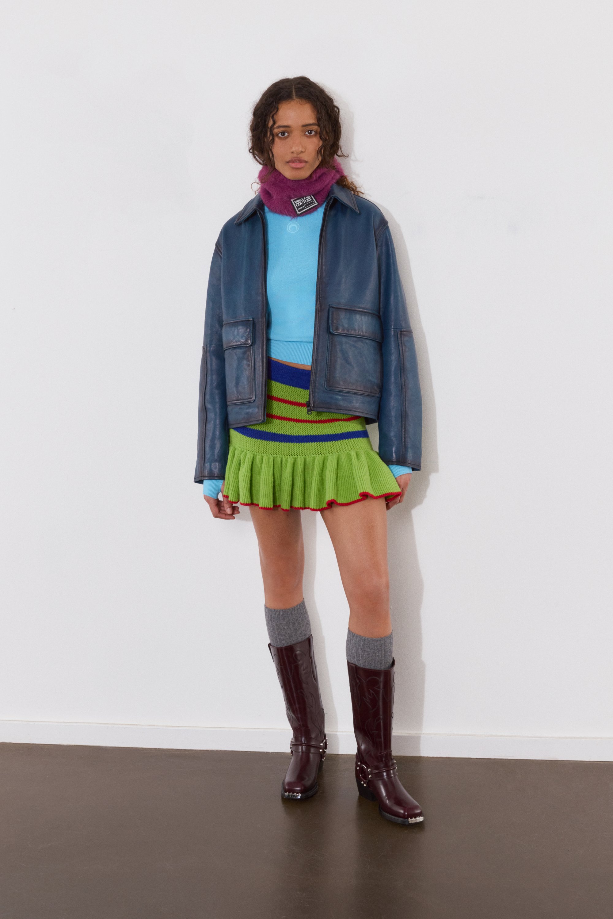 Una modelo se encuentra de pie frente a una pared blanca, vistiendo una blusa azul, una falda verde y una chaqueta de cuero, complementada con botas altas hasta la rodilla y calcetines grises.