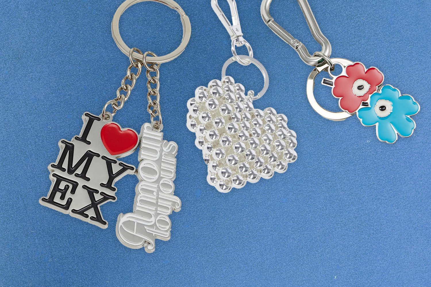 Llavero que presenta la frase "AMO A MI EX" en negro y rojo, un corazón transparente con cuentas elevadas, y dos encantos de flores de colores en rojo y azul.