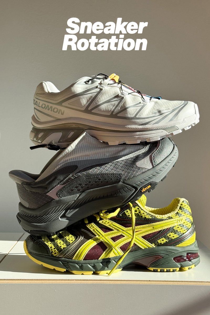 Tres zapatillas apiladas en una estantería: una Salomon gris en la parte superior, un zapato gris oscuro en el medio y una Asics negra/amarilla en la parte inferior.