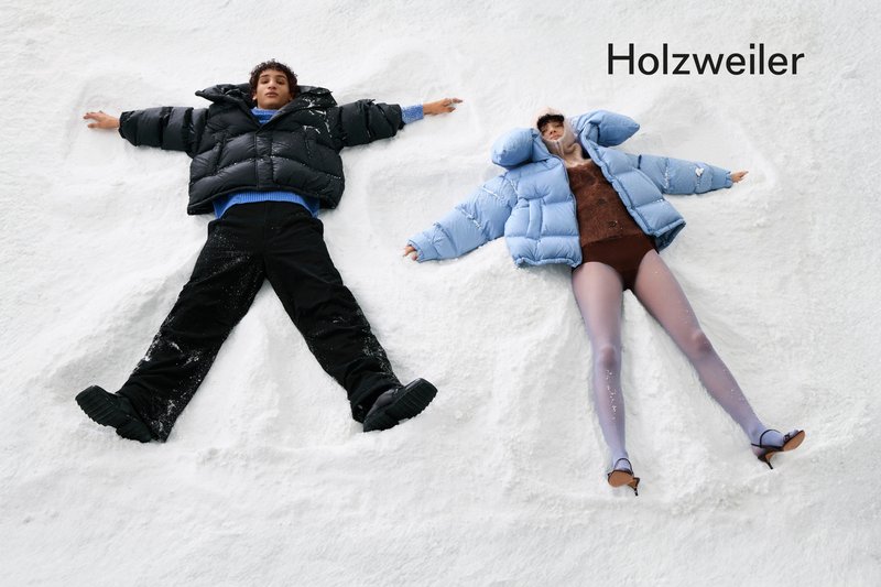 Due persone giacciono nella neve, creando angeli di neve. Una indossa un giubbotto imbottito nero e un maglione blu; l'altra indossa un giubbotto imbottito azzurro e collant trasparenti.