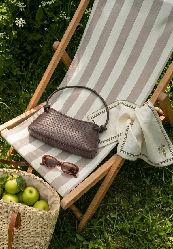 Sdraio a righe beige e bianca con borsa intrecciata marrone, occhiali da sole, panno bianco ricamato e un cestino di mele verdi sull’erba.