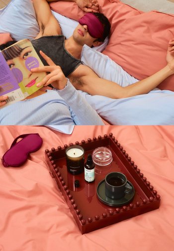 Un vassoio color granata contiene una candela, un contenitore di vetro, un contagocce per olio e una tazza scura con piattino. Una maschera per dormire rosa riposa su una biancheria da letto morbida e stratificata.