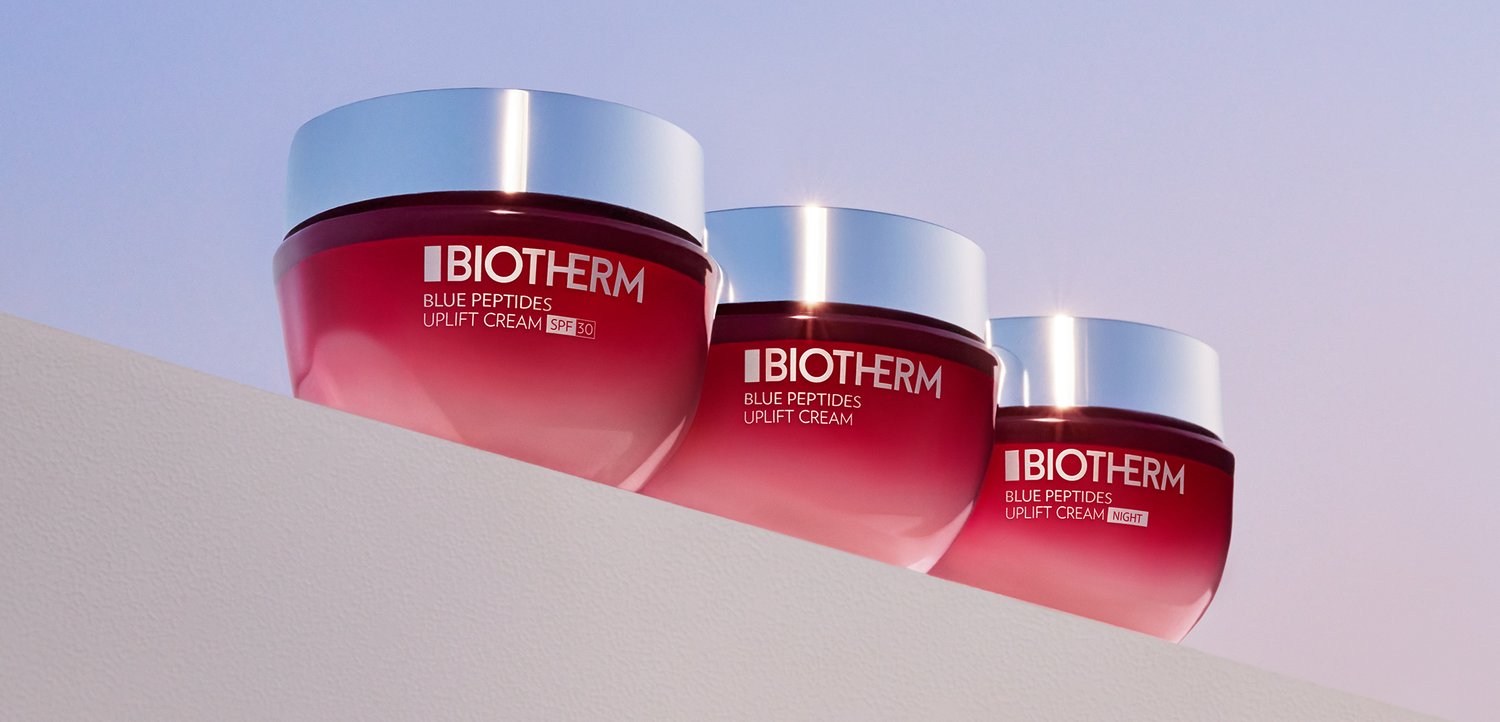 Trois pots de crème Biotherm Blue Peptides Uplift dans un emballage dégradé rouge, étiquetés pour un usage de jour et de nuit, sur un fond pastel doux.