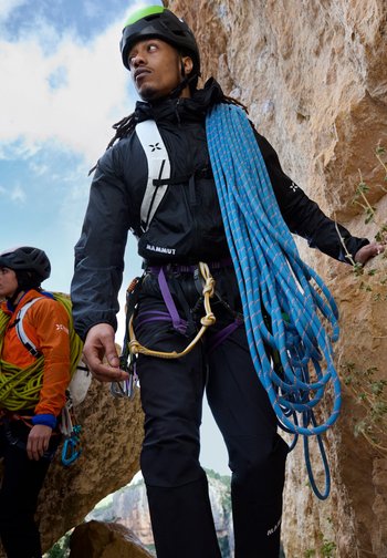 Two climbers wearing helmets and gear stand between rocky cliffs, with one carrying a coiled blue climbing rope over his shoulder.