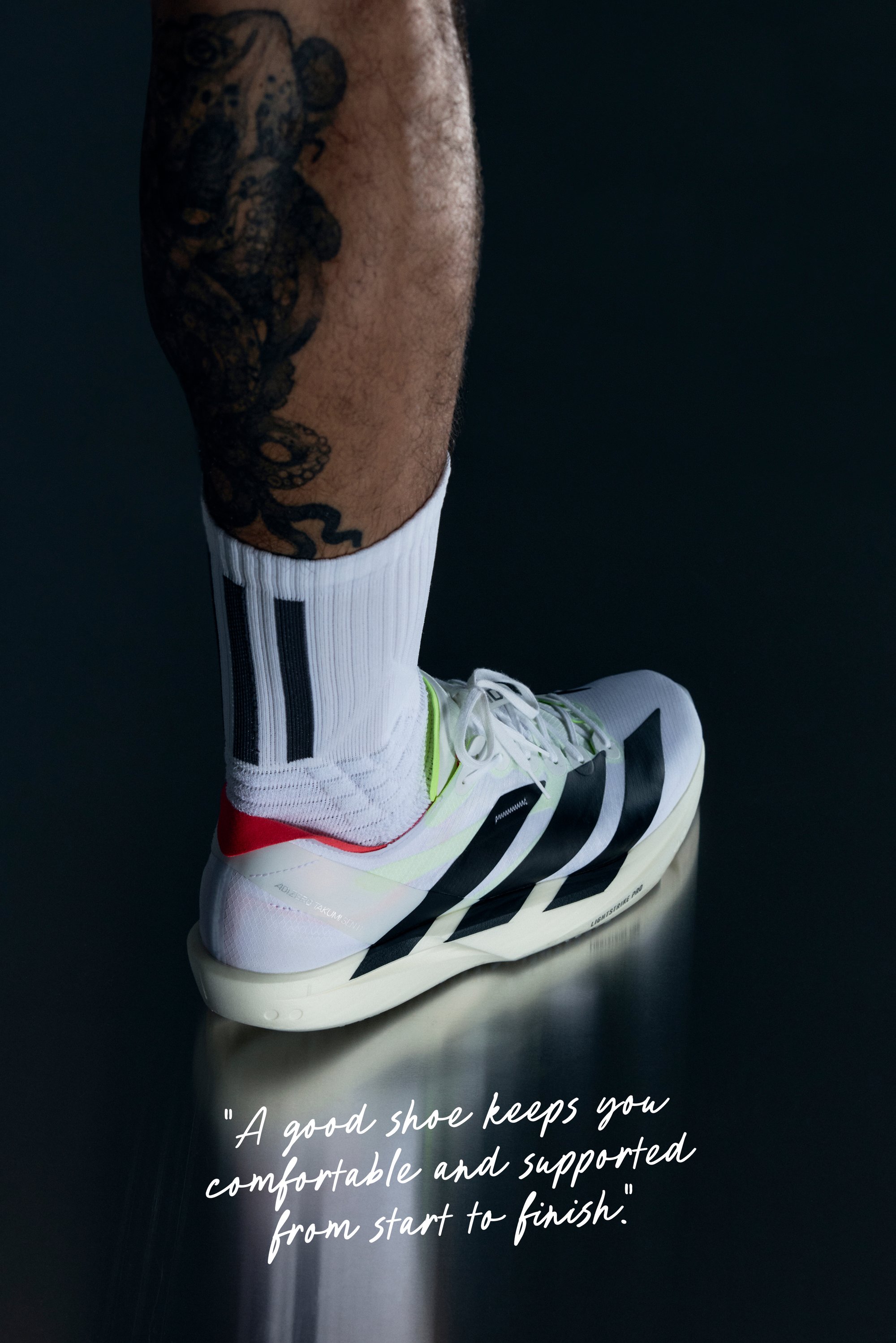 Bein, das einen weißen Sportschuh mit schwarzen Streifen und eine weiße Socke trägt, tätowierter Unterschenkel, stehend auf einer reflektierenden dunklen Oberfläche mit einem Zitat darunter.