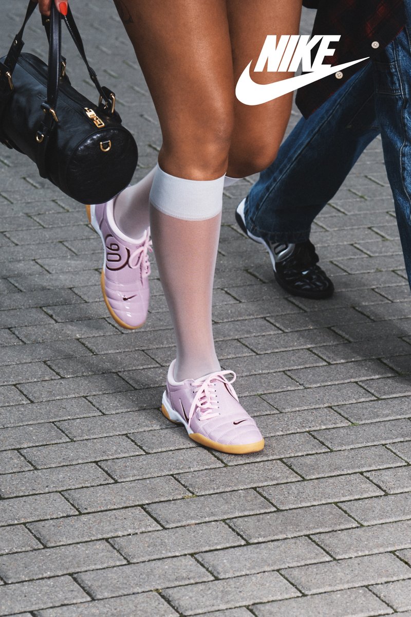 Zapatillas Nike rosas con cordones y acentos negros, llevadas con calcetas blancas hasta la rodilla. Un bolso negro y jeans de mezclilla son visibles en el fondo.