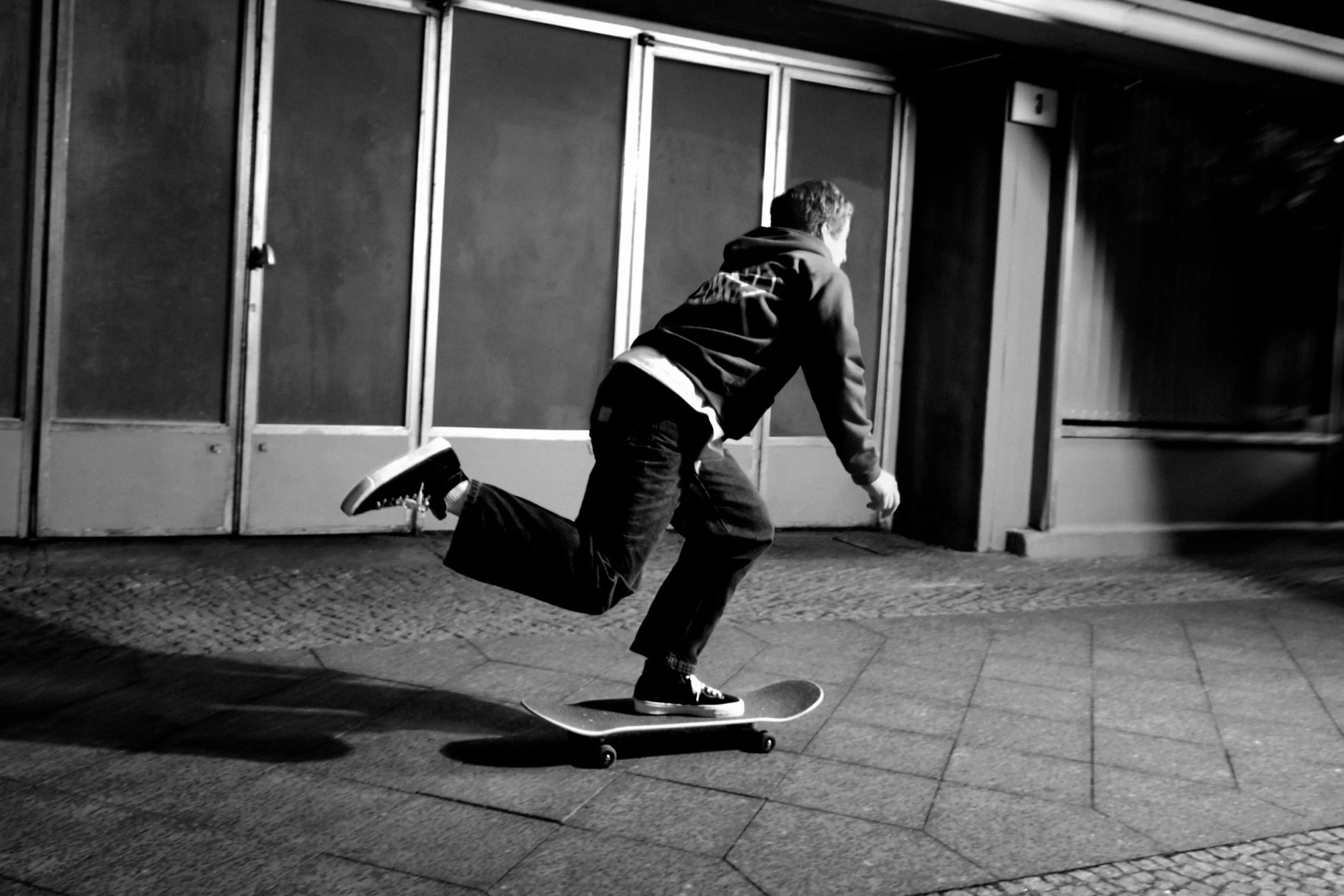 Una persona va in skateboard lungo una strada poco illuminata di notte, indossando un cappuccio e jeans, con un piede sollevato dalla tavola.