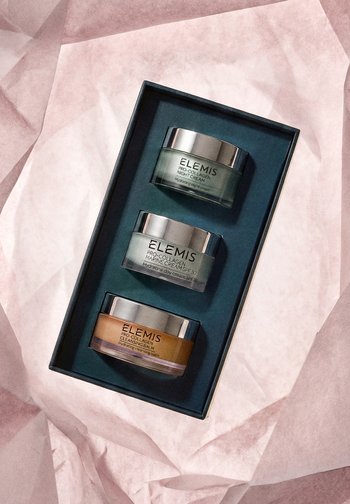 Trzy słoiczki kosmetyków Elemis Pro-Collagen — krem na noc, morski krem na dzień z filtrem SPF 30 oraz balsam oczyszczający — ułożone w czarnym pudełku na różowym papierze.