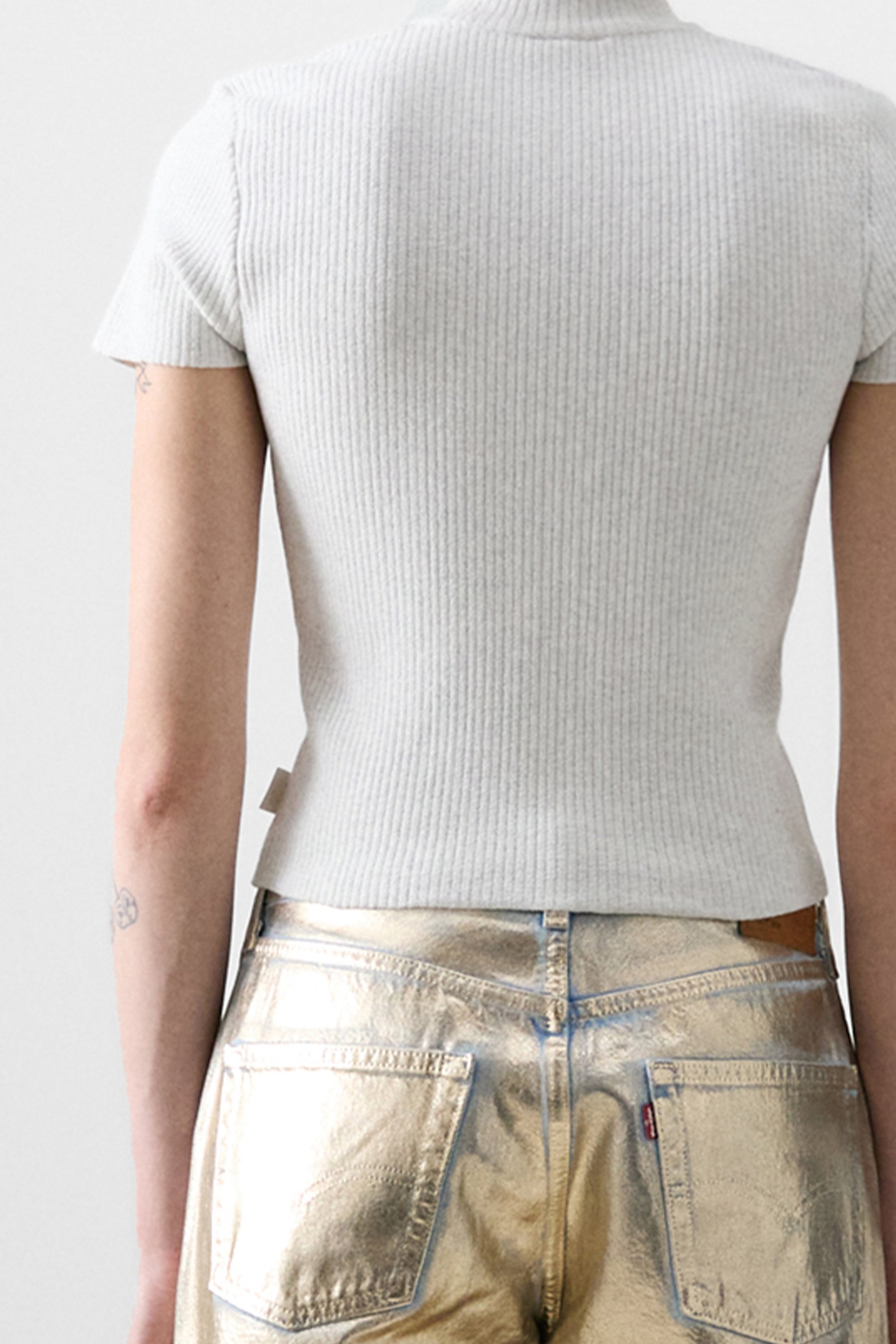 A person wears a fitted, ribbed light gray shirt and shiny gold pants, seen from the back, with tattoos visible on their arm.