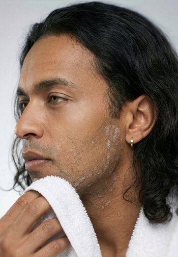 Uomo con capelli bagnati e orecchino che si asciuga il viso e il collo con un asciugamano bianco.