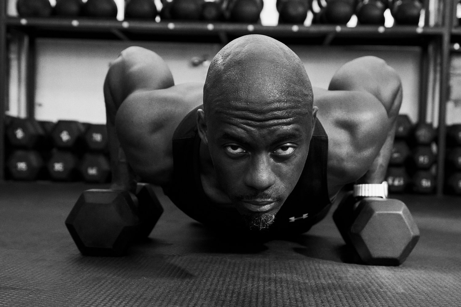 Svalnatá osoba vykonáva kľuky na telocvičnom koberci, čelom k fotografovi, s jednoručnými činkami vedľa seba a s činkami v pozadí.