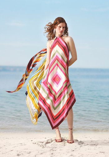 Vrouw draagt een halterjurk met veelkleurig geometrisch patroon en vloeiende stof, staand op een zandstrand bij een rustige zee.