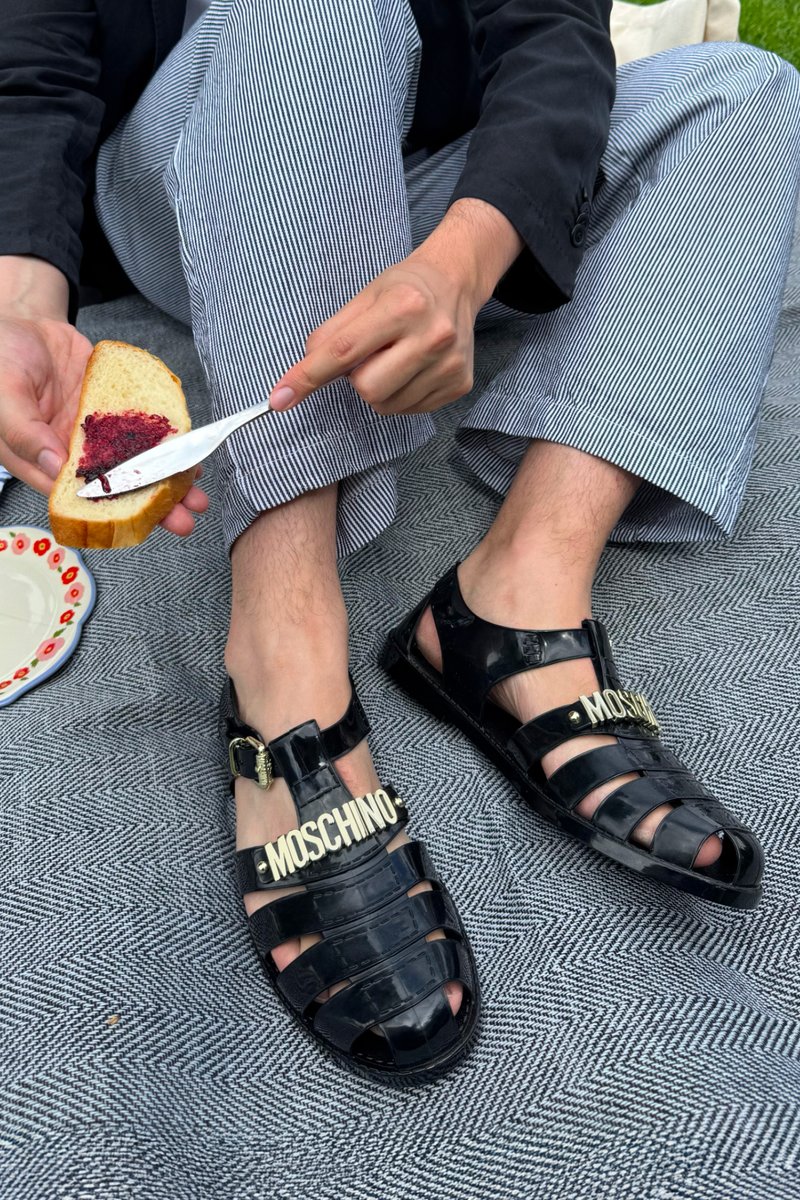 Infradito in gomma nera con design a intaglio, logo 'MOSCHINO' dorato e fibbia regolabile. Sono visibili dei pantaloni a righe e una fetta di pane spalmata.