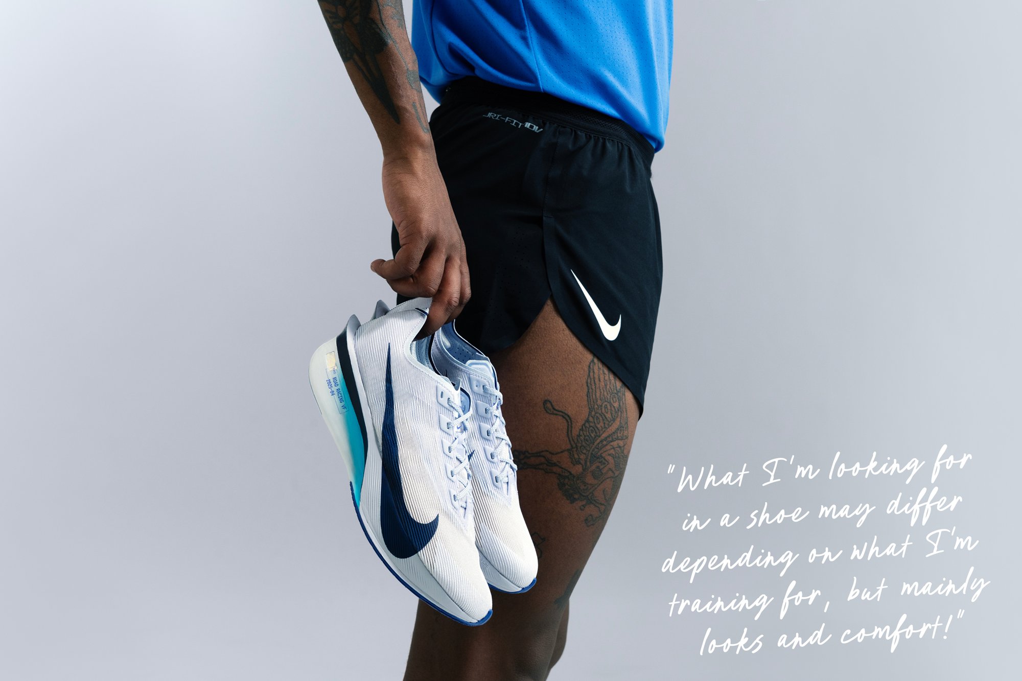 Weiße und marineblaue Nike Laufschuhe, die in einer Hand gehalten werden, mit einem strukturierten Design und dämpfender Sohle. Schwarze Sportshorts und ein blauer Oberteil sichtbar.