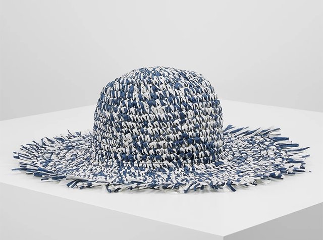 Sombrero de sol tejido de ala ancha con un patrón de rayas azules y blancas y bordes deshilachados, exhibido sobre una superficie blanca.