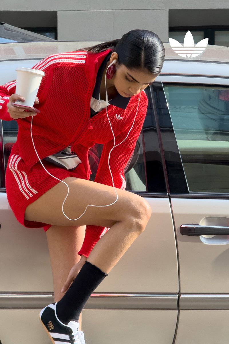 Een vrouw in een rode sportoutfit zit op de rand van een zilveren auto, houdt een koffiekop vast en draagt oordopjes terwijl ze gefocust naar haar telefoon kijkt.