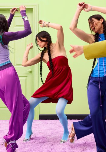 Trois personnes dansent avec énergie à l'intérieur sur un tapis violet, portant des vêtements colorés dont une robe rouge et des collants bleus.