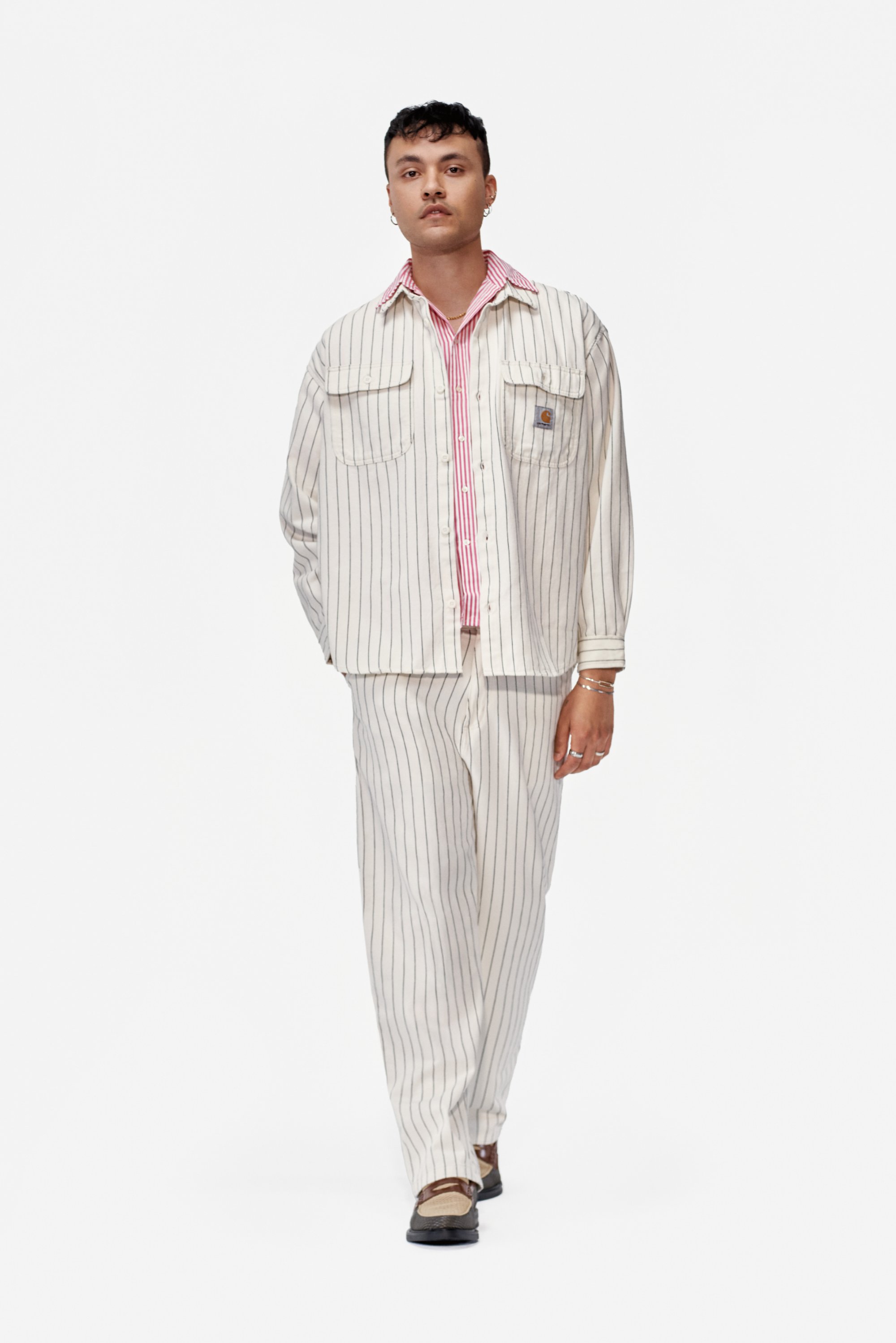 Un joven camina hacia la cámara, vistiendo un conjunto de rayas color crema con una camisa de cuello rojo y zapatos simples, exudando un estilo casual.
