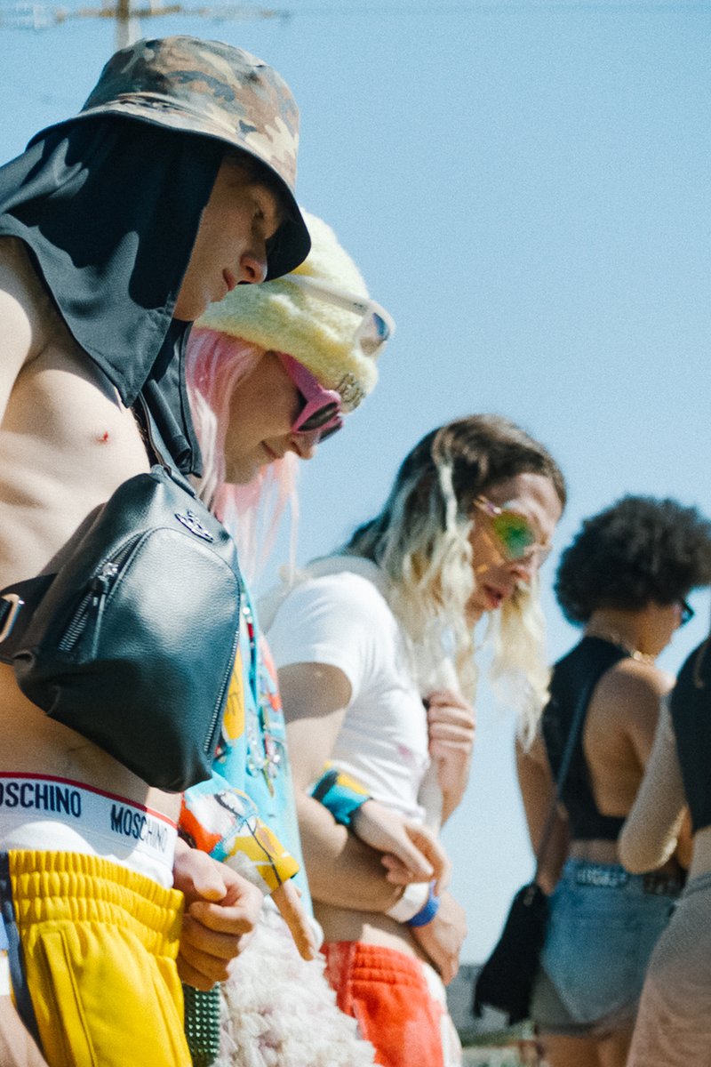 Een diverse groep individuen in stijlvolle outfits en accessoires wacht in een zonnige omgeving, waarbij levendige kleuren en unieke modekeuzes worden getoond.