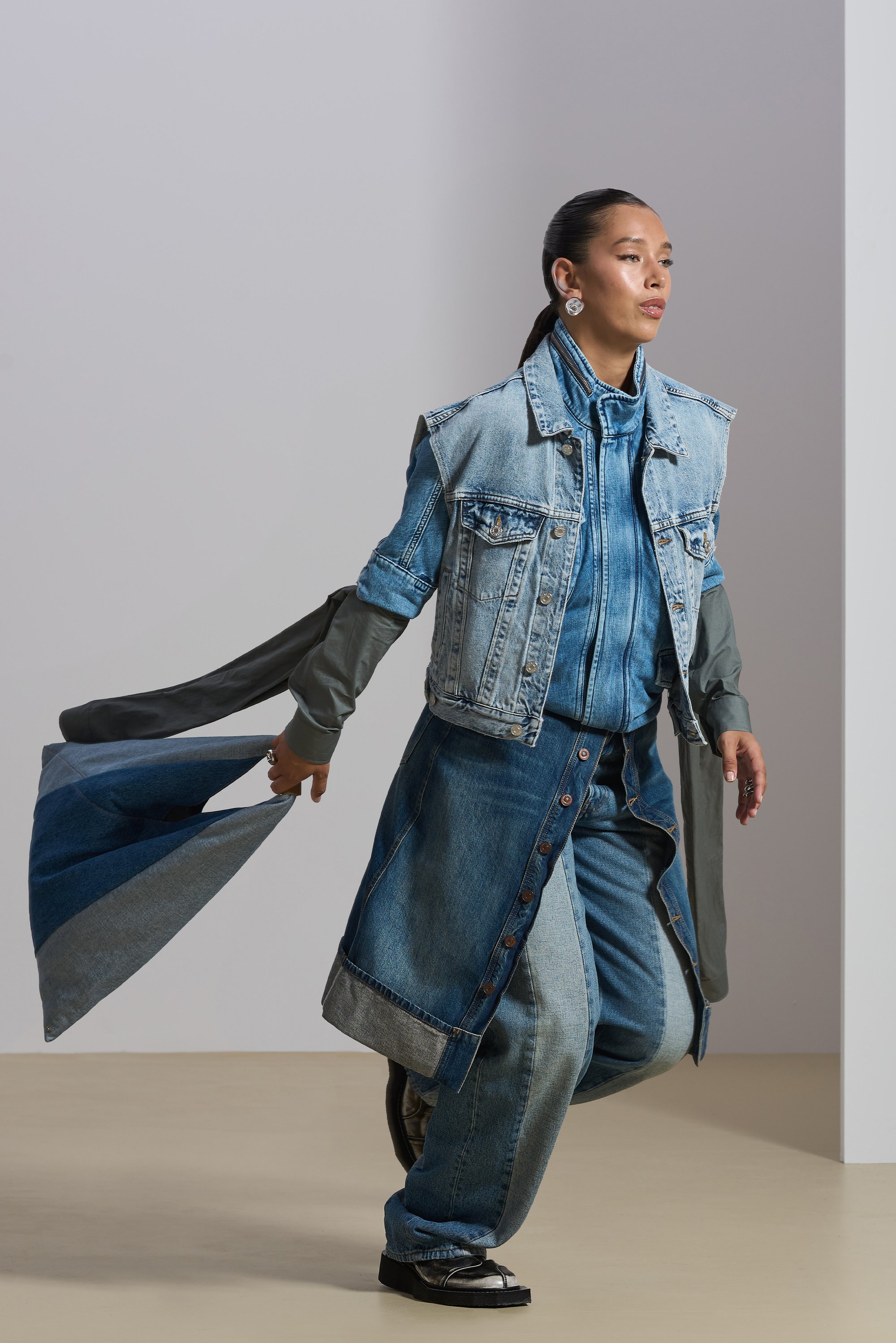 A person in a layered denim outfit confidently walks, holding a flowing fabric. The background is minimalistic, emphasizing their fashion.