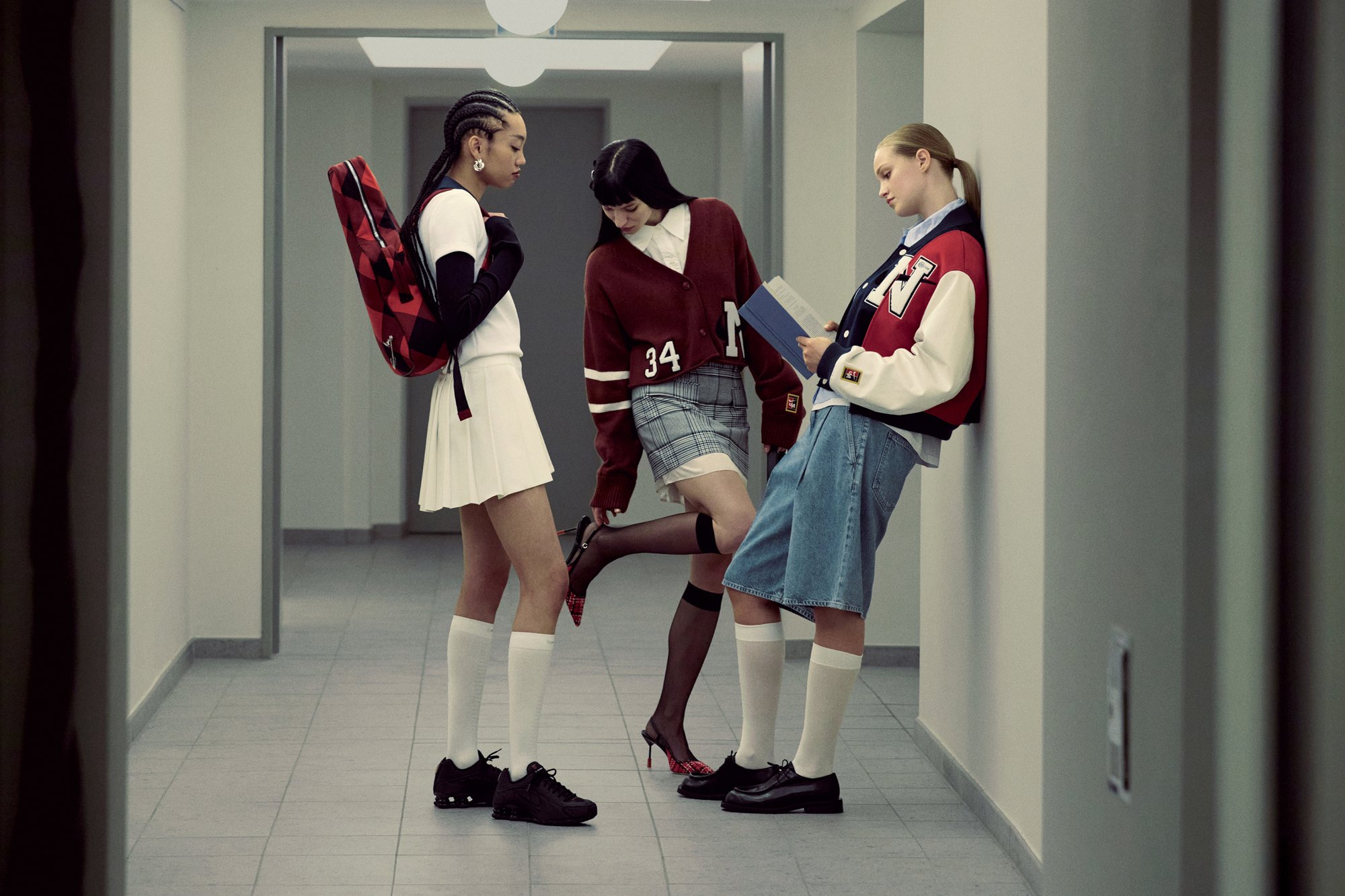 Tre giovani donne in abbigliamento alla moda interagiscono in un moderno corridoio, mostrando stili vari: sportivo, preppy e casual.