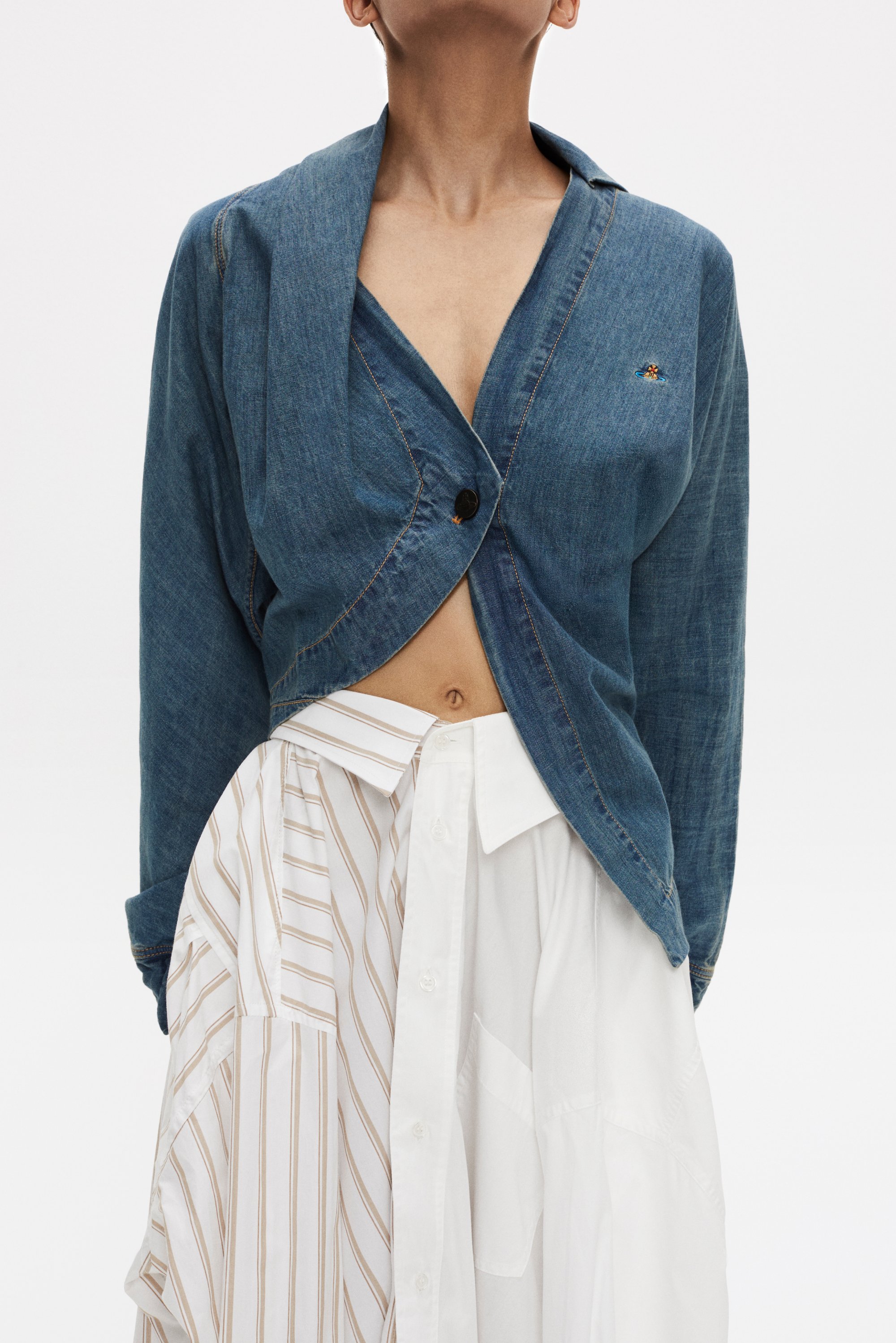 Modella che indossa una giacca di jeans aperta sopra una camicia bianca a righe verticali, che mette in mostra un outfit moderno e rilassato su uno sfondo neutro.